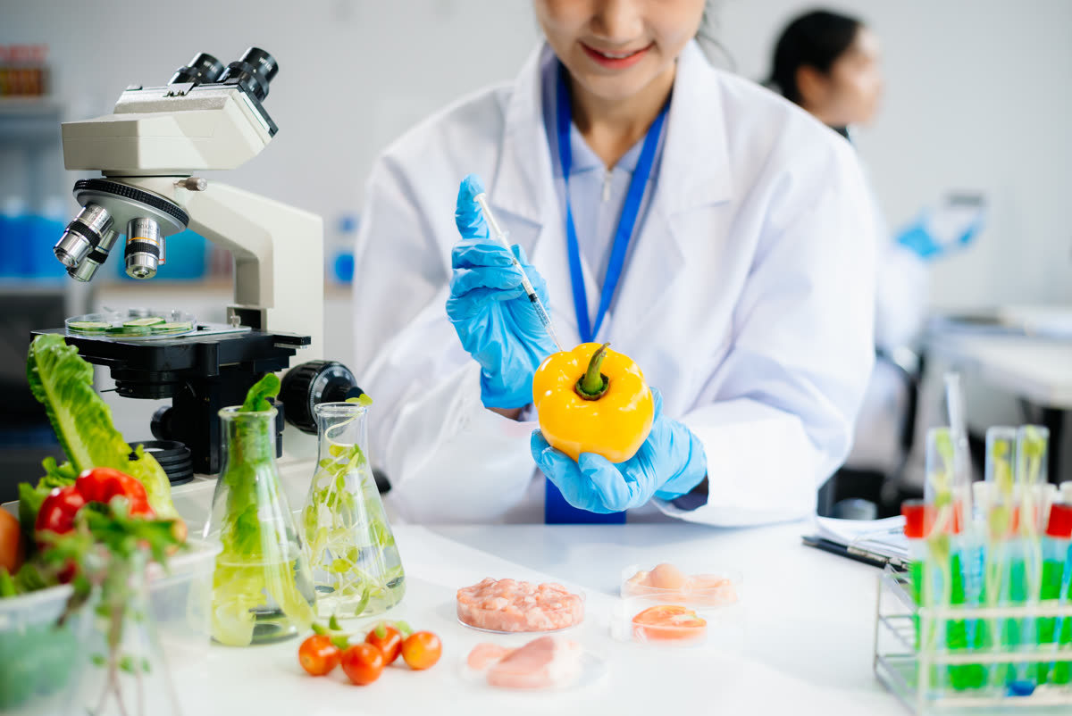 Analista de laboratorio con bata y guantes inyectando una muestra en un pimiento, junto a microscopio, matraces y cajas Petri con muestras de alimentos.