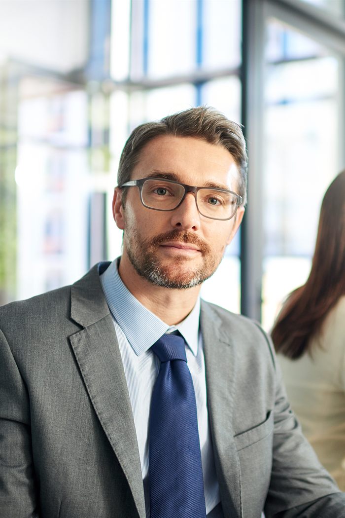 Hombre de traje gris y lentes en una sala de juntas, con colegas reunidos al fondo.