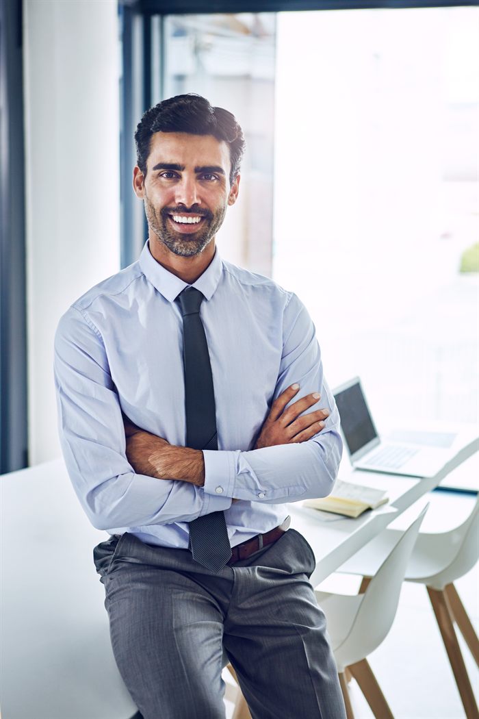 Profesional de camisa y corbata apoyado en un escritorio, con una computadora portátil al fondo.